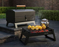 Two black outdoor grills on a stone patio, with one larger charcoal grill sitting on a raised stone platform and a smaller folding grill in front cooking corn on a stone surface. A silver teapot sits on the smaller grill.