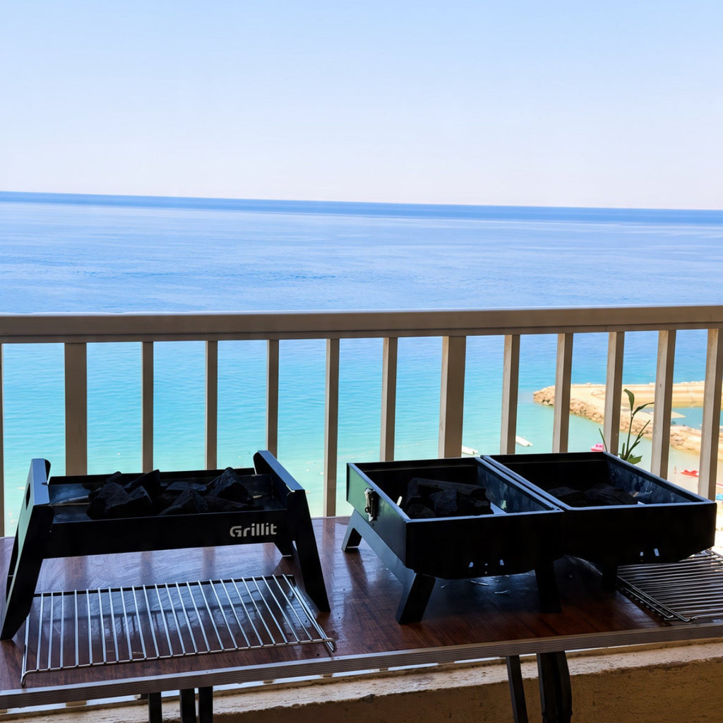 Two black portable outdoor grills on a folding table on a balcony overlooking a scenic blue ocean and sky.