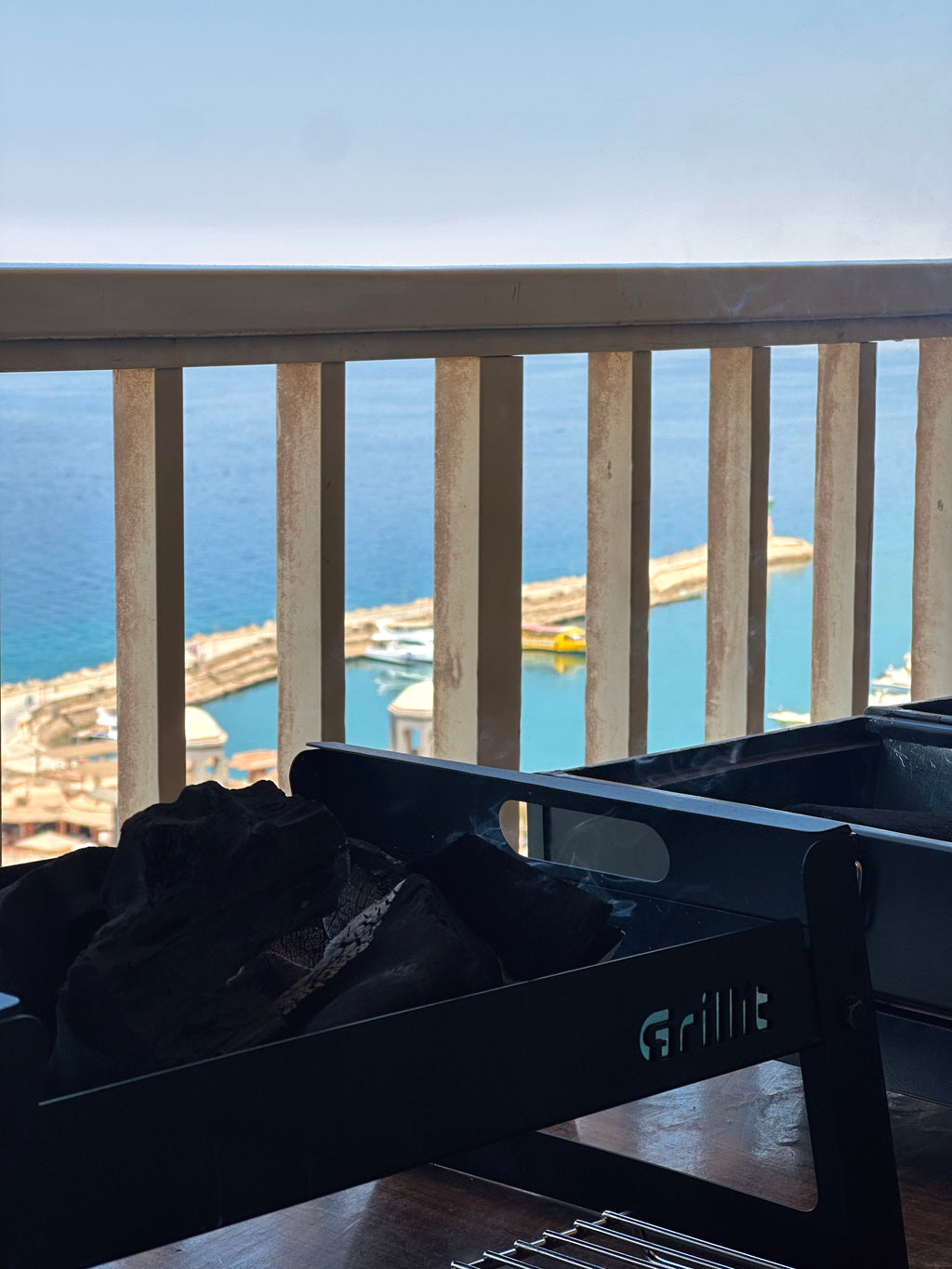 A black metal outdoor grill on a balcony overlooking a harbor and blue sea, with the brand name Grillit visible on the side.