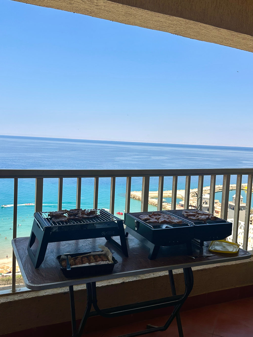 Two portable black charcoal grills cooking food on a folding table on a balcony, with a clear view of a blue ocean and beach under a bright sky.