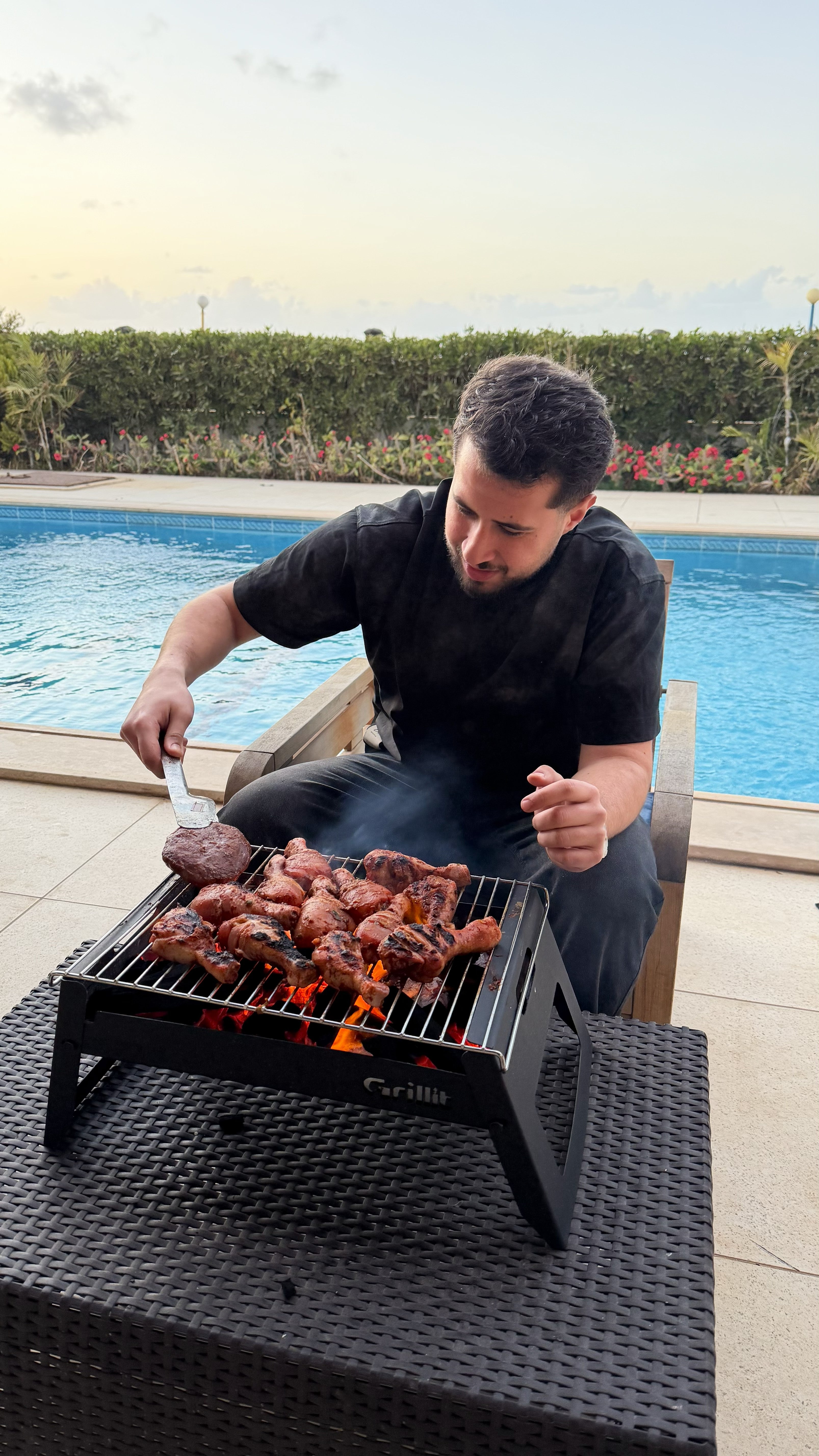Man grilling food on a small barbecue grill by a poolside.